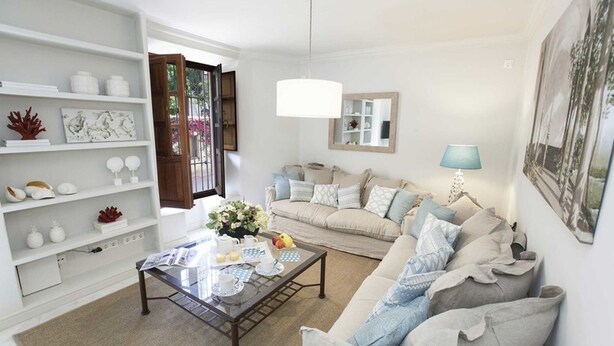 Una sala de estar con un sofá color beige, una mesa de centro de vidrio y estanterías blancas con objetos decorativos.