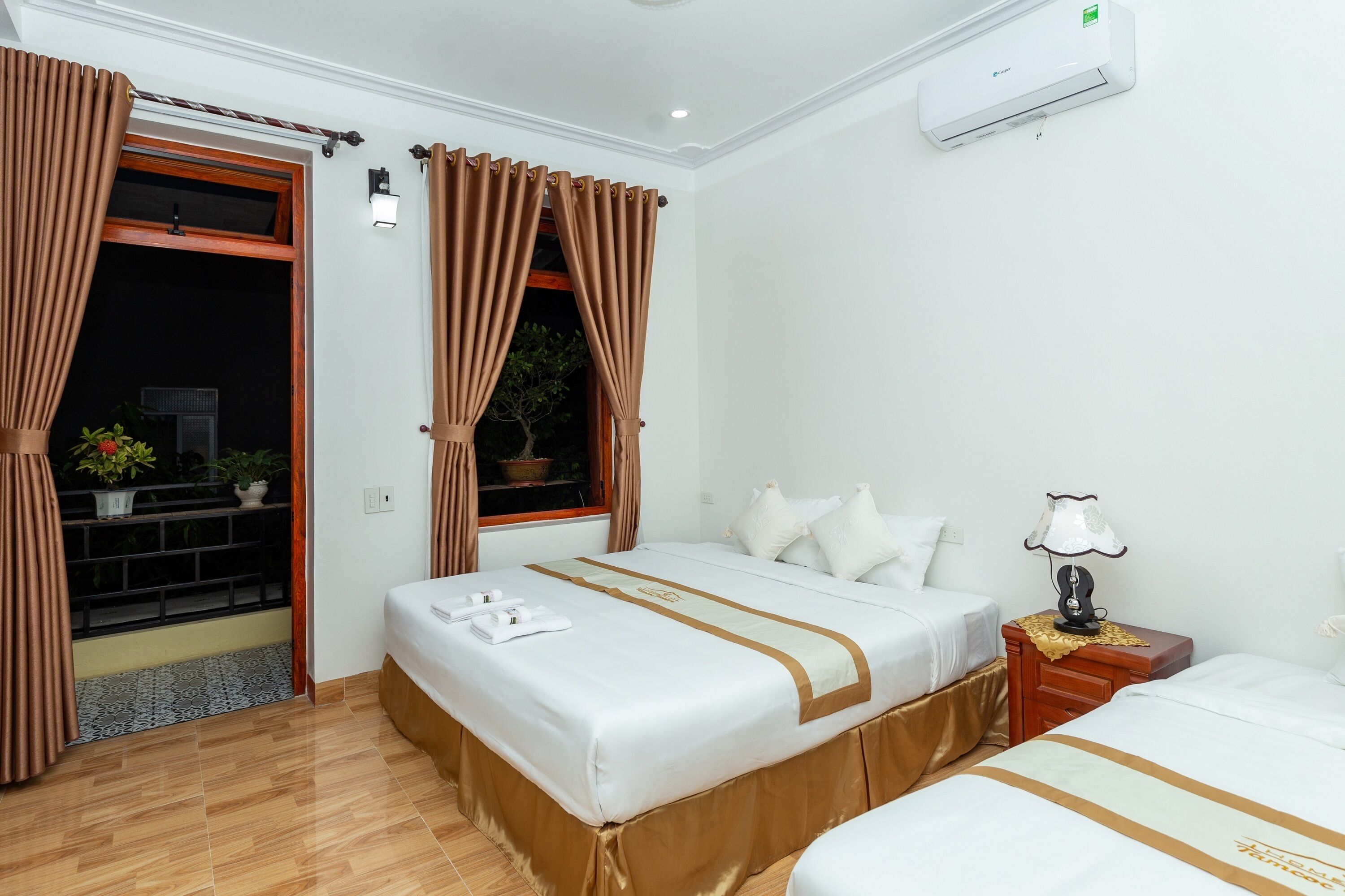 A hotel room with a bed, a bedside table with a lamp, a window with curtains, and an air conditioning unit.