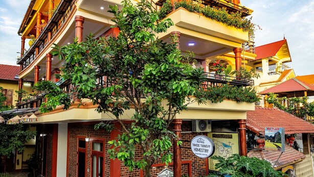 A multi-story building with balconies, a sign for 'Mai Place Homestay', and a large tree in front.
