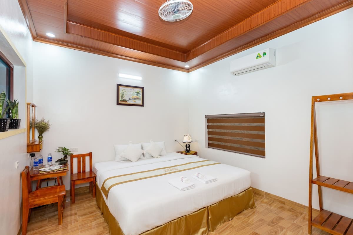 Une chambre d’hôtel avec un plafond en bois, un lit avec une literie blanche, un bureau avec une chaise, une petite table avec une lampe et une fenêtre avec des stores.