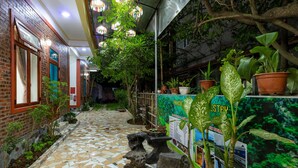 A narrow, tiled pathway leading to a building with brick walls and wooden doors. There are potted plants along the sides and a metal fence.