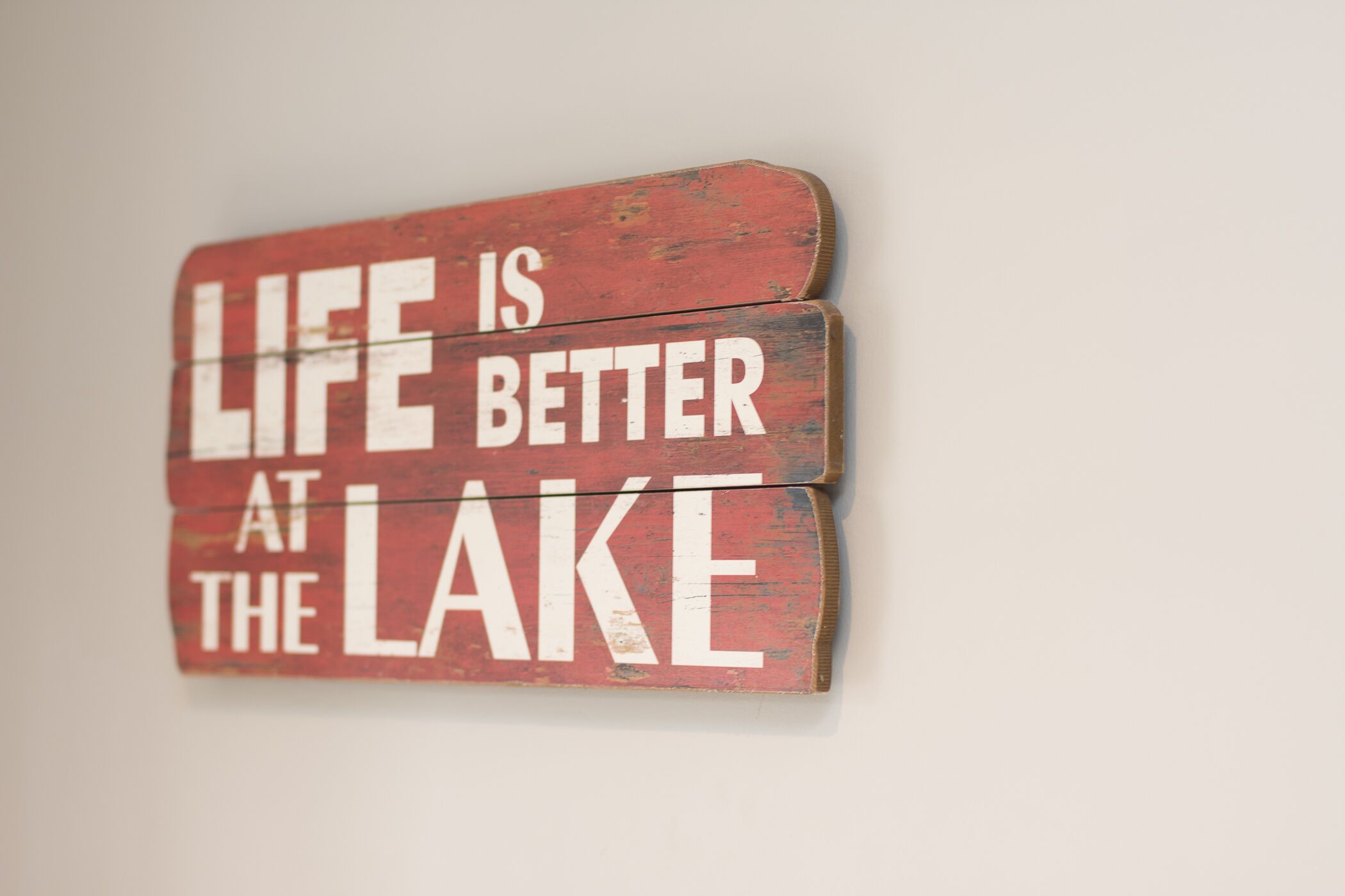 A wooden sign with the text "LIFE IS BETTER AT THE LAKE" mounted on a wall.
