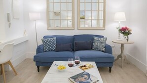 Un salón con un sofá azul, dos ventanas, una mesa de centro blanca con comida y bebidas, y una mesita con una lámpara y flores.