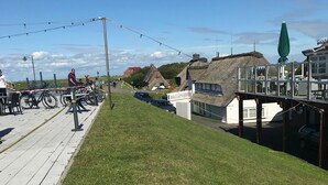 Ein Rasenstück mit einer Holzterrasse, einem Gebäude mit Strohdach und einem gläsernen Anbau mit Tischen und Stühlen.