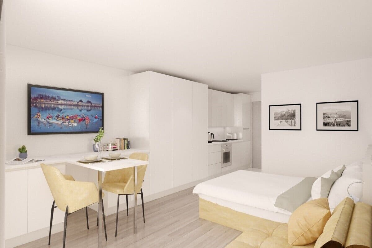 A modern hotel room with a bed, a desk, and chairs. There are framed pictures on the wall and a window with a view of a body of water.