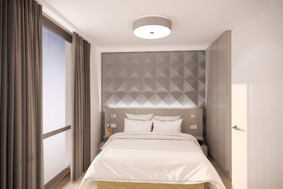 A modern hotel room with a large bed, a geometric-patterned headboard, a bedside table with a lamp, and a view through a large window with curtains.