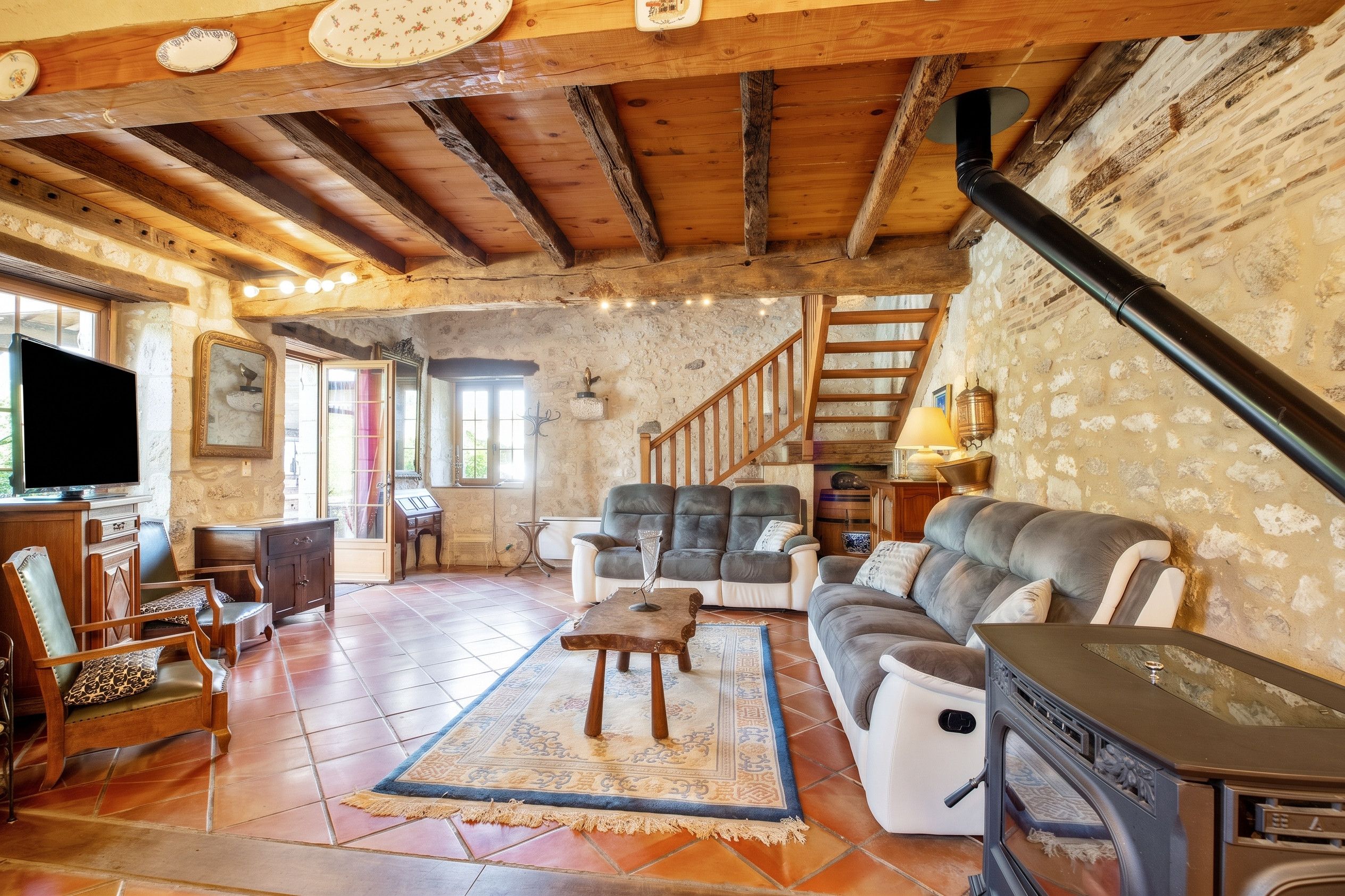 A rustic living room with a fireplace, wooden beams, stone walls, and a staircase.