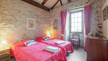 Ein Zimmer mit zwei Betten, einem Holfnachtkästchen, einem Stuhl, einem Holzkasten und einem Fenster mit Vorhängen.