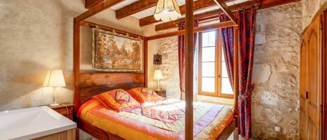 Un lit à baldaquin surmonté d’une courtepointe aux couleurs vives, un mur en pierre, un plafond en bois et un tableau accroché au-dessus du lit.