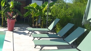 Een zwembadgedeelte met ligbedden en een grote parasol, omringd door potplanten en een gebouw op de achtergrond.