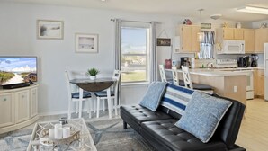 Una sala de estar con un sofá de cuero negro, una mesa de centro y un televisor. Se observa un comedor con una mesa y sillas. La cocina es visible, con gabinetes y electrodomésticos.