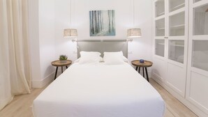 Una cama bien hecha con ropa blanca, dos mesitas de noche con lámparas, un cuadro en la pared y un amplio armario con puertas de vidrio.