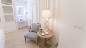 Habitación de hotel con un sillón gris, una mesa redonda de madera con una lámpara, una pequeña planta en maceta y una mesita de noche blanca con un teléfono y cargador.