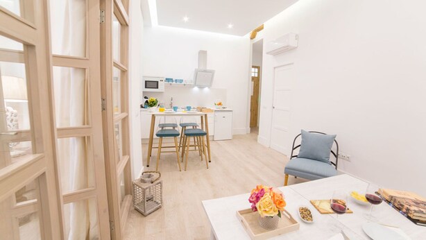 Una cocina moderna y luminosa con armarios blancos, microondas y un refrigerador pequeño. Cuenta con un comedor con una mesa blanca, sillas azules y una mesita auxiliar con un florero.