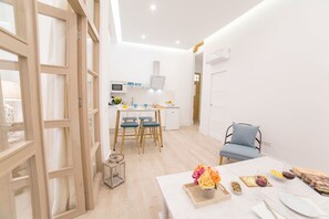 A bright, modern kitchen with white cabinets, a microwave, and a small refrigerator. There is a dining area with a white table, blue chairs, and a small side table with a vase of flowers. - GR Suites Boutique Catedral AP-A (Málaga)