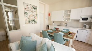 Un salon moderne avec un canapé blanc et des coussins turquoise, un coin repas avec une table en bois dressée pour quatre personnes, et une cuisine avec des meubles blancs et un four à micro-ondes.