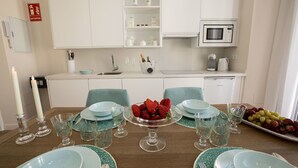 Une cuisine moderne avec des meubles blancs, un four à micro-ondes et un évier. On y trouve une table à manger dressée avec des assiettes, des verres et un plateau de fruits.