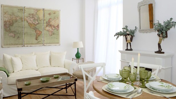 Quarto com sofá branco, mesa de centro de madeira, mesa de jantar posta com copos verdes, mapa na parede e lareira com duas velas.