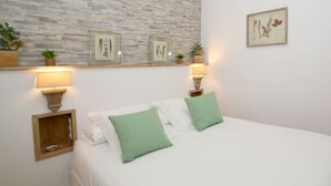 Uma cama bem arrumada com almofadas brancas e verdes, duas mesas de cabeceira com abajures e gravuras botânicas emolduradas na parede.