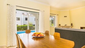 A dining area with a wooden table, chairs, and a fruit bowl. There is a window with curtains, a view of a pool, and a sofa in the background. - Jadlyn Yellow Villa, Falésia Beach, Albufeira, AC, WiFi, Swimming Pool !New! (Albufeira)