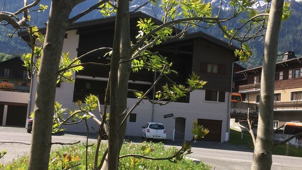 Un edificio de dos pisos con un automóvil estacionado adelante, rodeado de árboles y con una montaña al fondo.