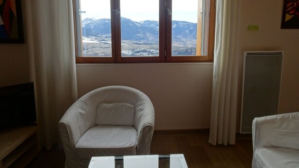 Une pièce dotée d’une grande fenêtre donnant sur les montagnes, d’un fauteuil blanc, d’une petite table et d’un radiateur.