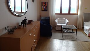 Une pièce comprenant une commode en bois, un miroir rond, une chaise bleue, un fauteuil blanc, une table basse en verre et une vue sur la mer, visible à travers de grandes fenêtres.
