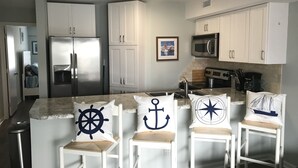 Cocina moderna con armarios blancos, electrodomésticos de acero inoxidable y una isla central con taburetes y almohadones con motivos náuticos.