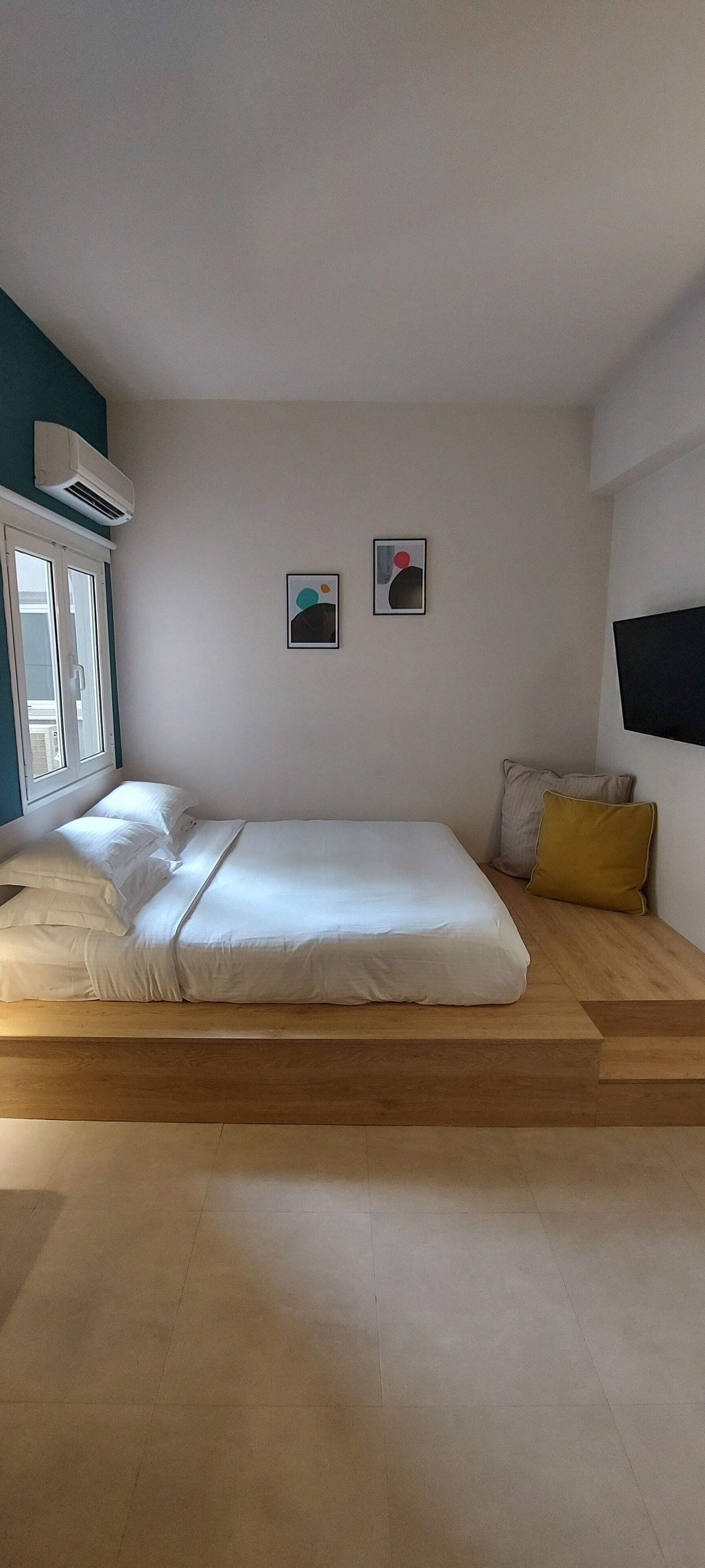 A bedroom with a bed, a window, an air conditioner, and a wall-mounted TV.