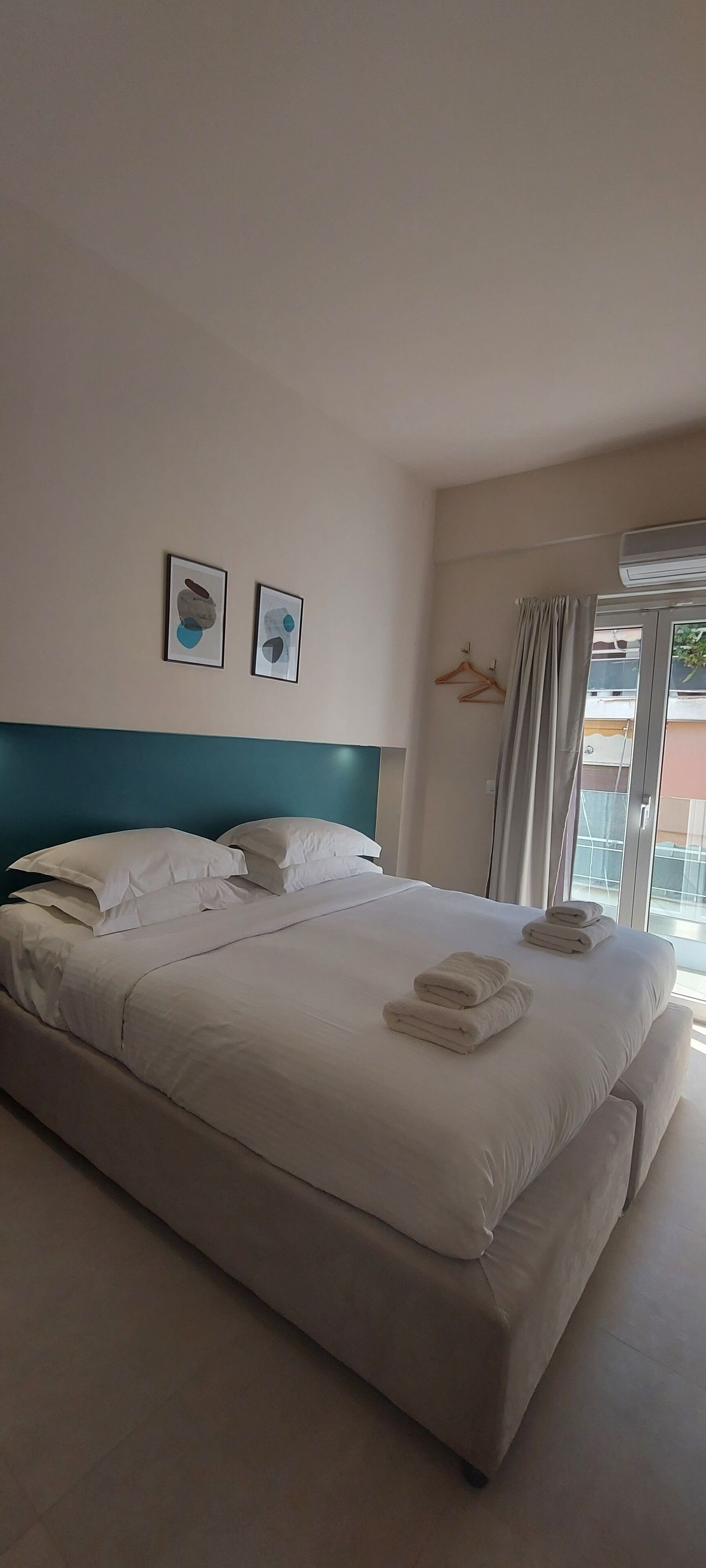 Une chambre d’hôtel avec un grand lit, une tête de lit de couleur turquoise, deux œuvres d’art encadrées, une fenêtre avec des rideaux et un balcon avec vue.
