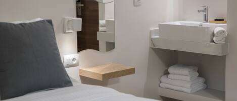 A modern hotel room with a bed, a bedside table with a lamp, a built-in shelf with towels, a mirror, and a wall-mounted light switch.