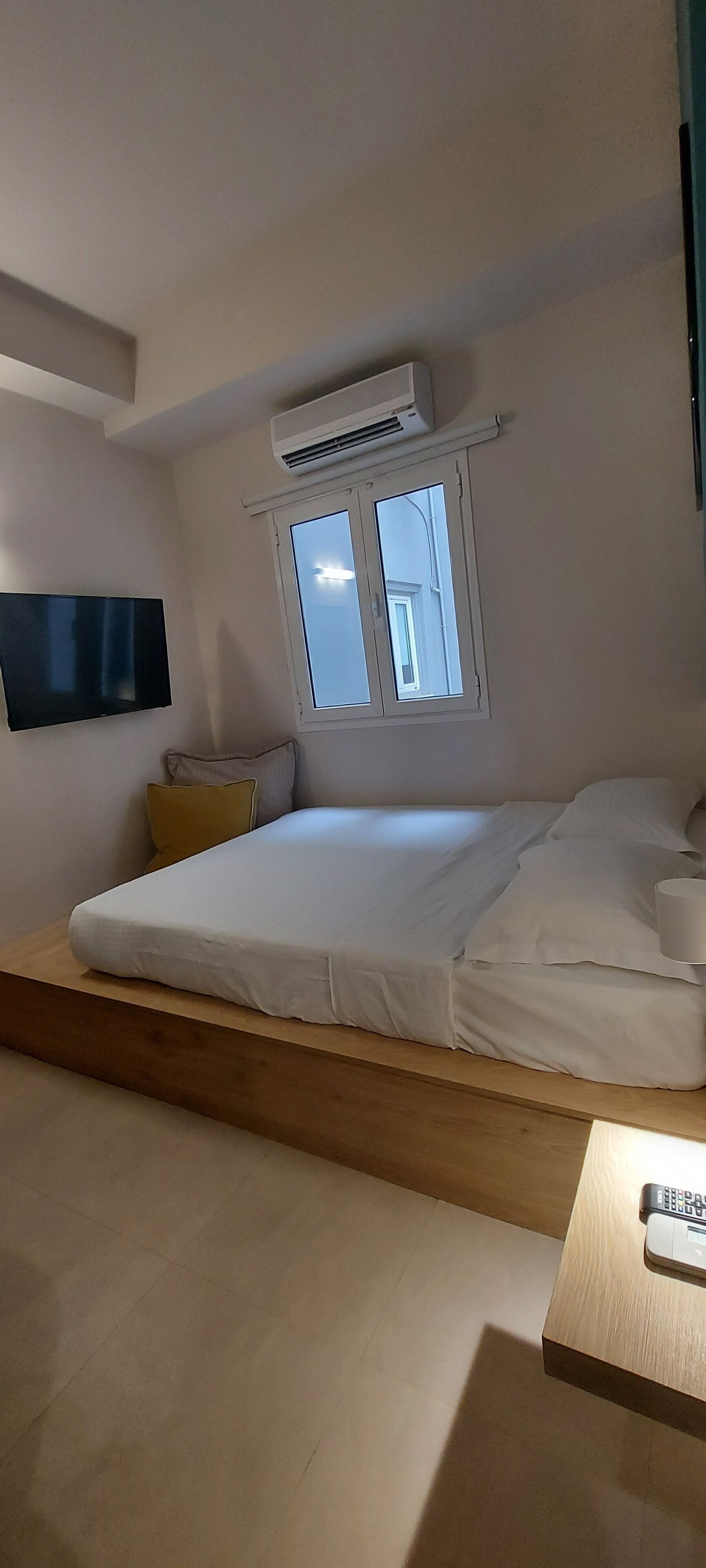 A bedroom with a bed, a window, an air conditioning unit, a television, and a remote control on a wooden table.