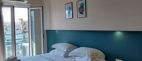 A modern hotel room with a large bed, a bedside table with a phone, and a view of the city through a large window.