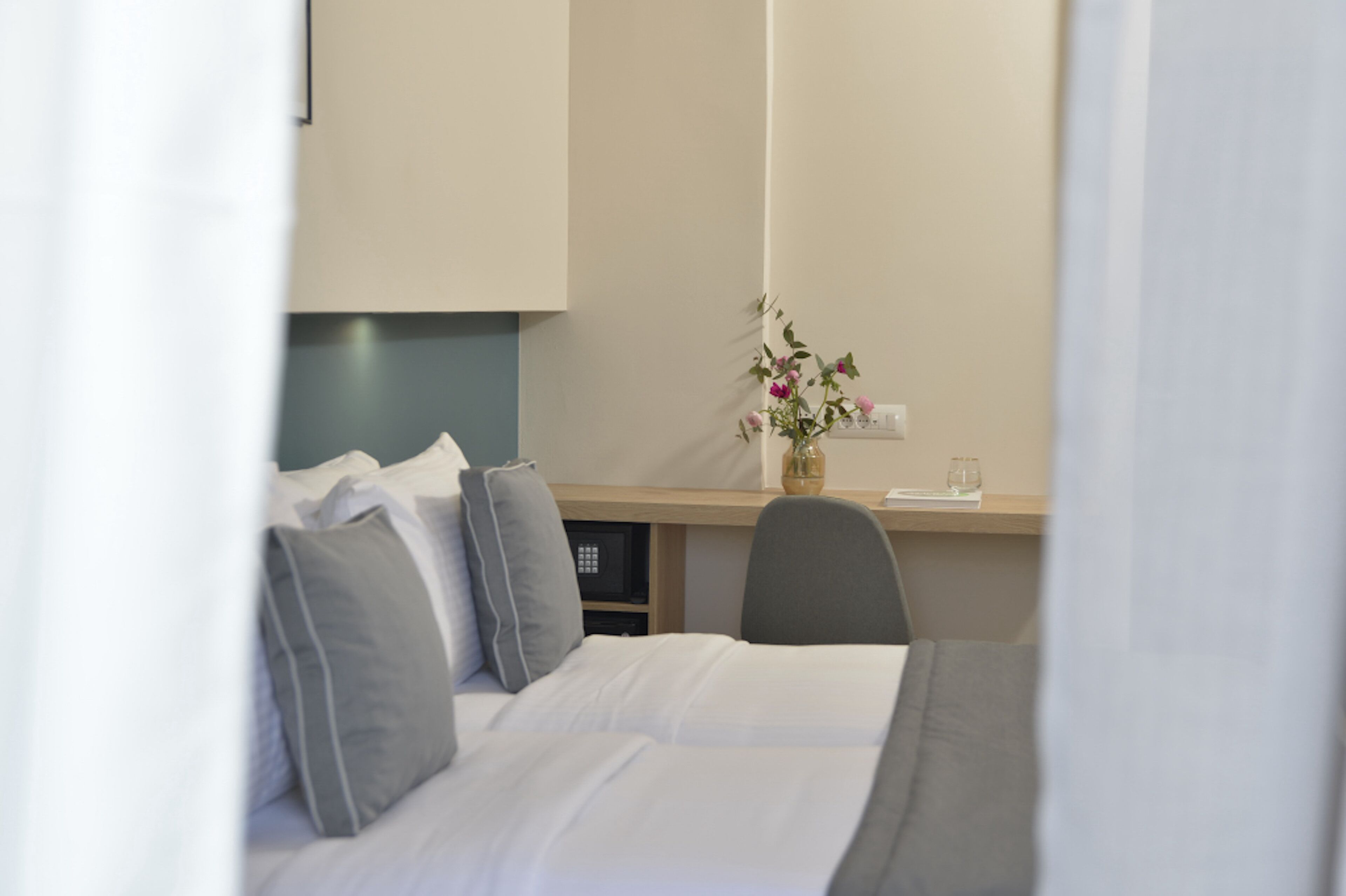 A hotel room with a bed, a desk, a chair, a vase with flowers, and a small table with a glass.