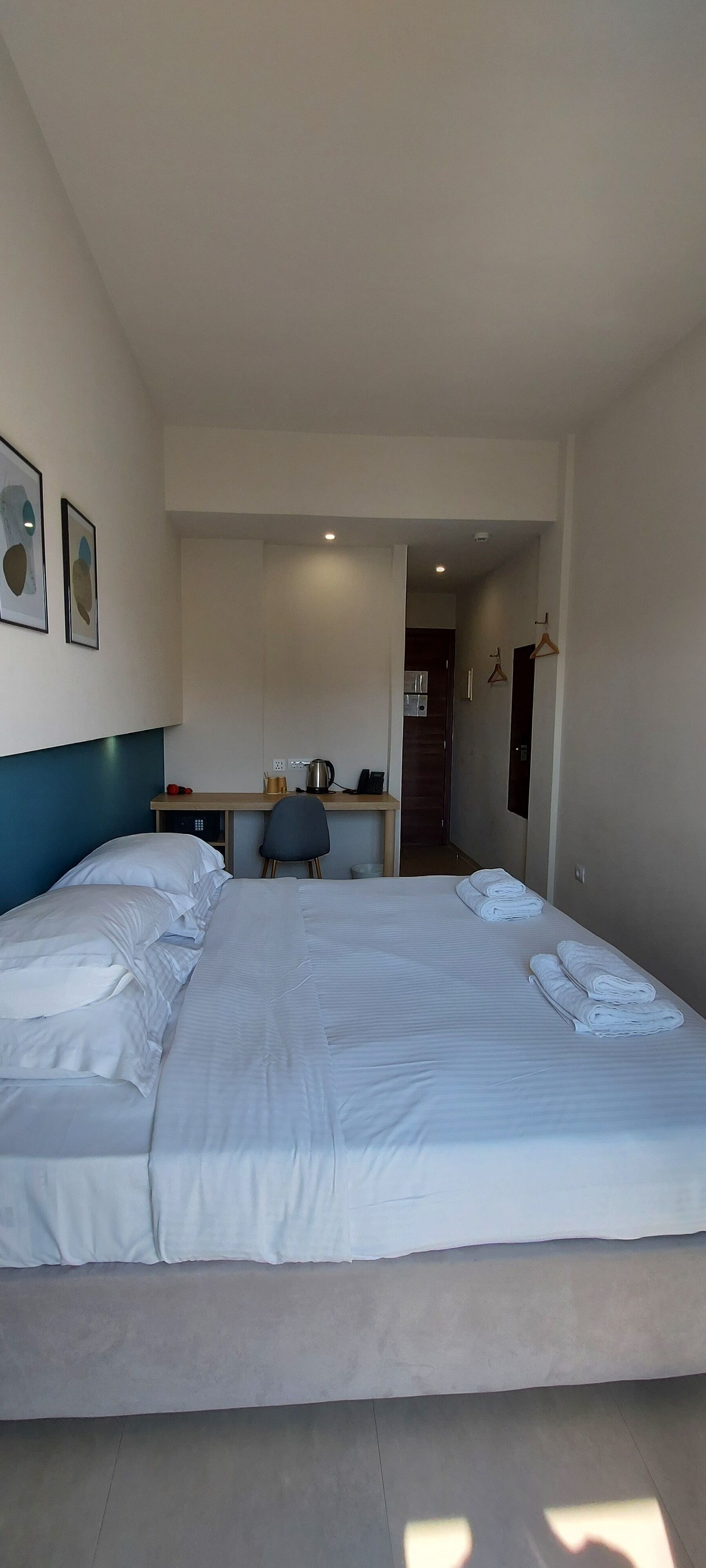 A hotel room with a large bed, a desk with a chair, and a kitchenette in the background.