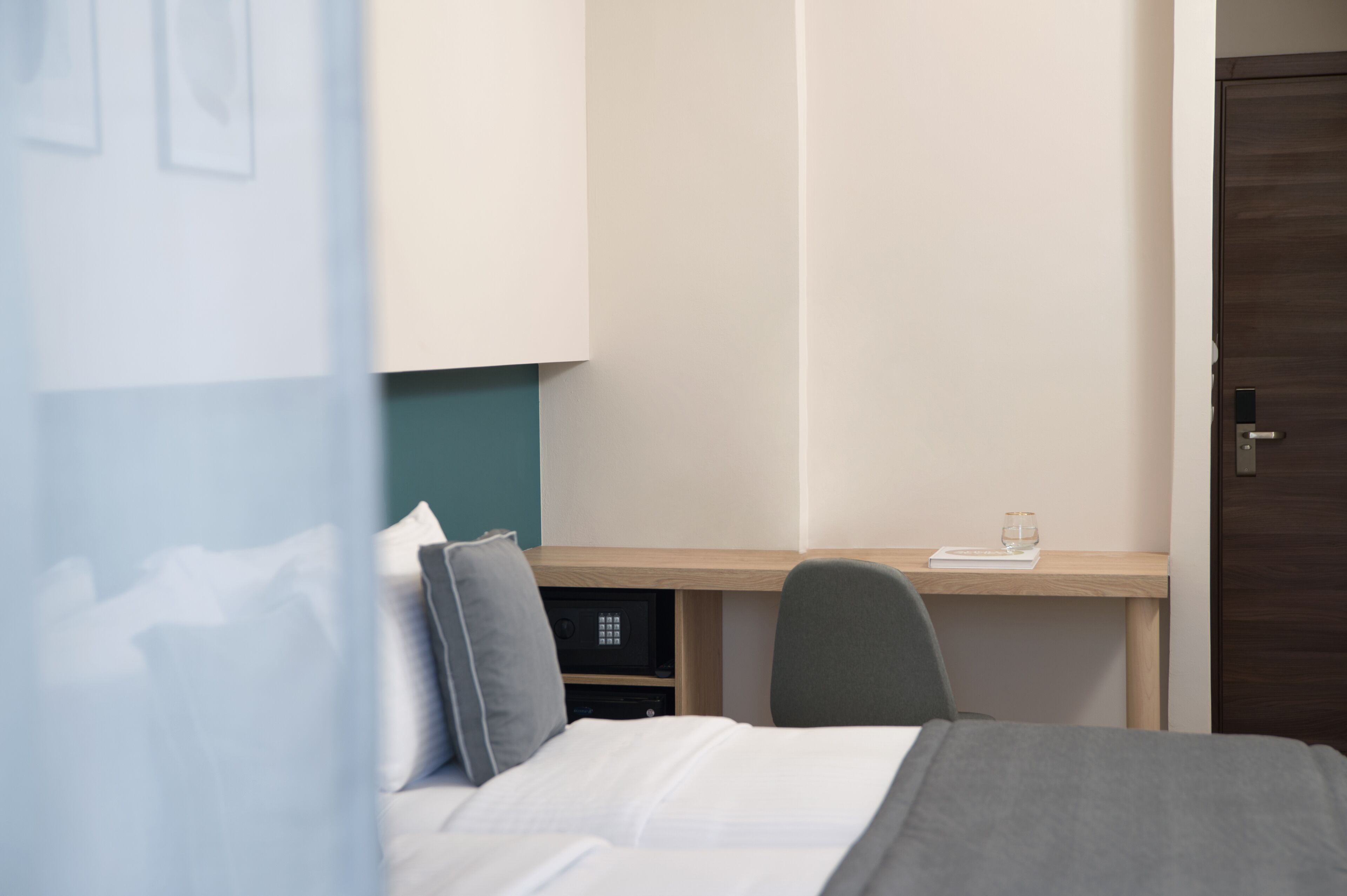 Une chambre d’hôtel avec un lit, un bureau et une chaise. On trouve une petite table sur le bureau, avec un verre et un livre dessus.