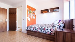 Une chambre avec un plancher en bois, un lit recouvert d’une courtepointe à motifs, une étagère fixée au mur avec des livres et un tableau d’affichage avec des notes.