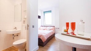 Une salle de bain moderne avec des toilettes, un lavabo et un miroir. La pièce est dotée d’un plancher en bois et d’un lit près d’une fenêtre.