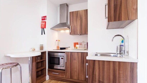 Une cuisine compacte avec des meubles en bois, un four encastré, un évier en acier inoxydable et un petit tabouret de bar.