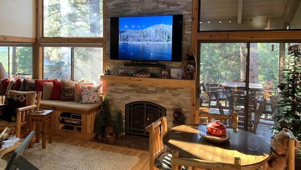 A cozy cabin interior with a stone fireplace, a television mounted above it, and a large window offering a view of the surrounding forest. - Northstar Mountain charm & luxury, near shuttle, 10 minutes to slopes, sleeps 6, (Truckee)