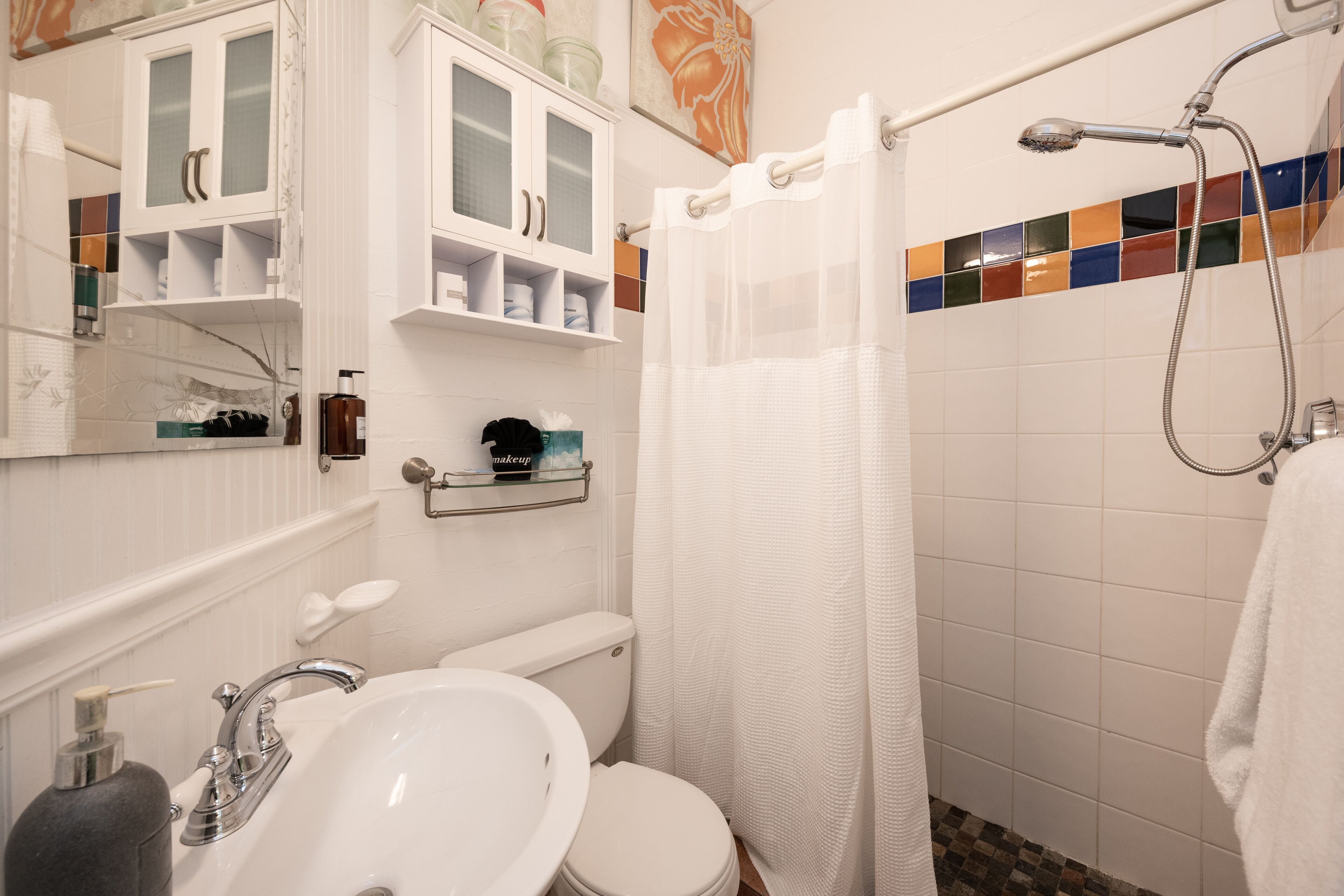 Un baño con lavamanos, inodoro y ducha blancos. Hay armarios sobre el lavamanos y una cortina de ducha.
