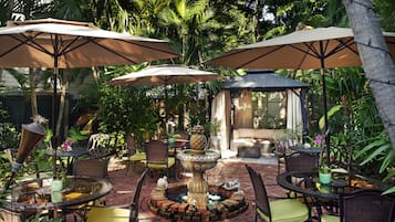 Eine Terrassengruppe mit runden Tischen, Stühlen und Sonnenschirmen, umgeben von üppiger Bepflanzung und einer Zierbrunnen.