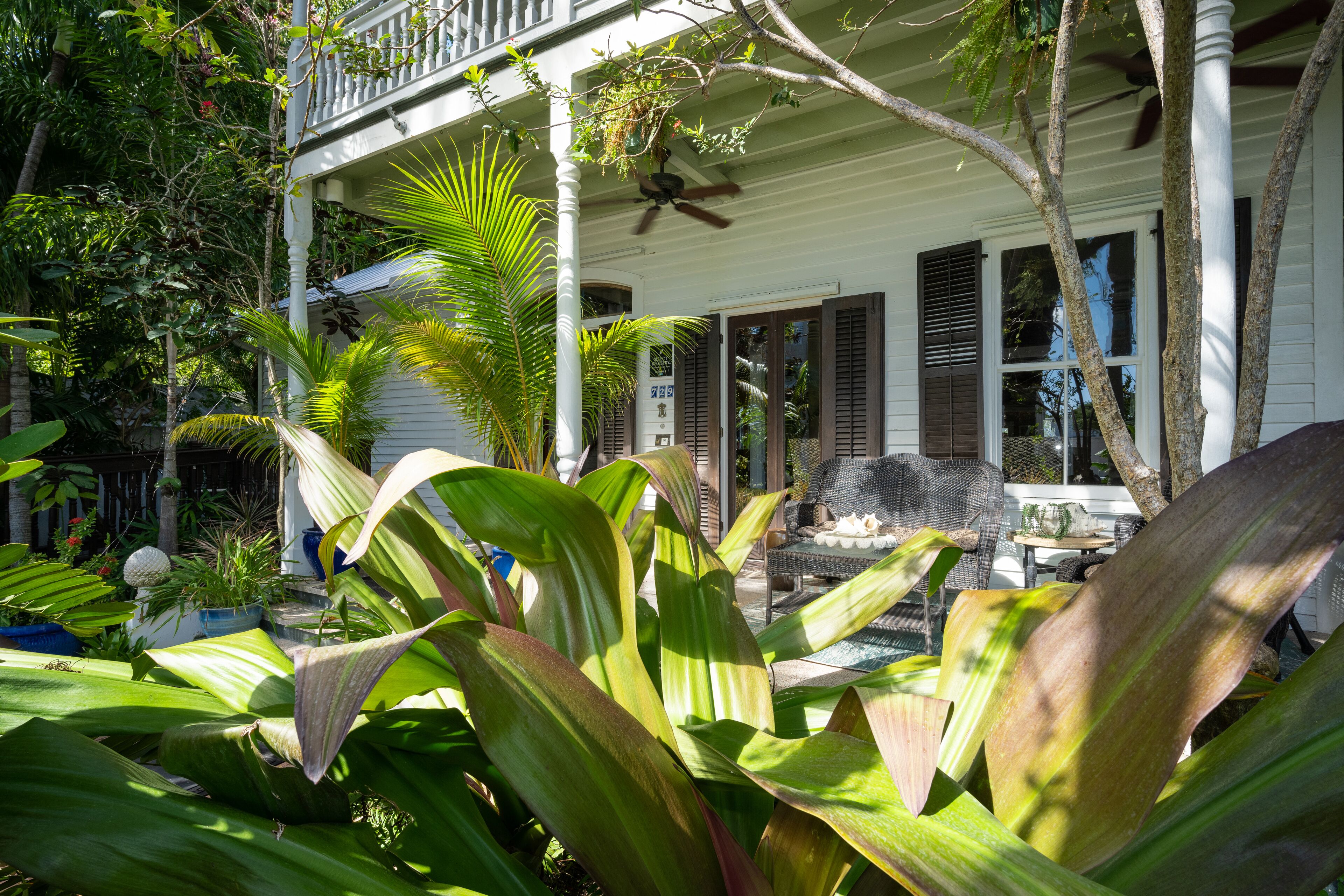 A two-story house with a balcony, surrounded by lush tropical plants.