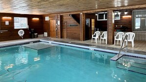 An indoor swimming pool with wooden walls and ceiling, a diving board, and white plastic chairs around the pool.