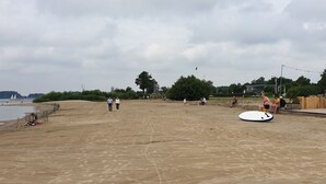 Een zandstrand met enkele mensen die wandelen, een zeilboot in de verte en een groot wit surfboard in het zand.