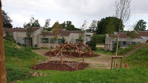 Een speeltuin met klimconstructies en een zandpad dat naar een woongebied met huizen leidt.