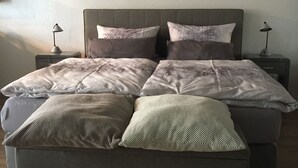 A bed with a grey upholstered headboard, multiple pillows, and a blanket. There are two bedside tables with lamps, and a painting of flowers on the wall. - Apartment mit Burgblick im Grünen, Familie Held (Burgthann)