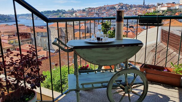 Ein Balkon mit einem antiken Wagen, Blumentöpfen und Blick auf eine Stadtlandschaft mit Terrakotta-Dächern.