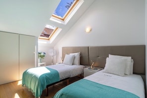 A bedroom with two beds, a skylight, a bedside table with a lamp, and a wall-mounted fan.