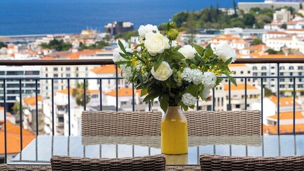 A balcony with wicker furniture, a glass table, and a yellow vase with white flowers. - Santa Luzia Funchal View | 2br pool, ac & parking (Funchal)